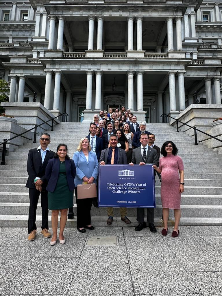 photo of a group of about 25 women and men standing on steps of a white-grey building that has 2 stories visible with pairs of pillars over the entrance. 3 people hold a large sign with logo of The White House and text 'Celebrating OSTP's Year of Open Science Recognition Challenge Winners dated September 19, 2024'