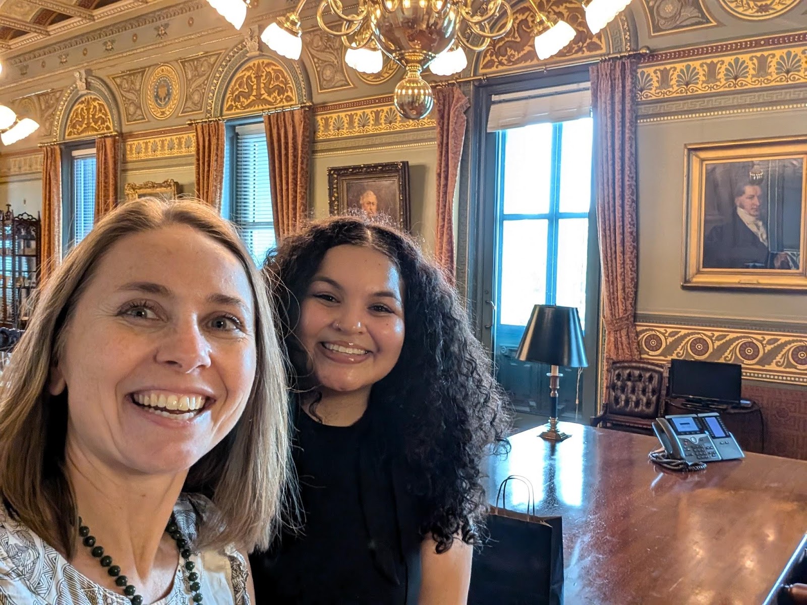 selfie of a white woman and a Black woman smiling standing under a chandelier and in front of an ornate wall with gilt accents with 3 large portraits of men and 3 large windows